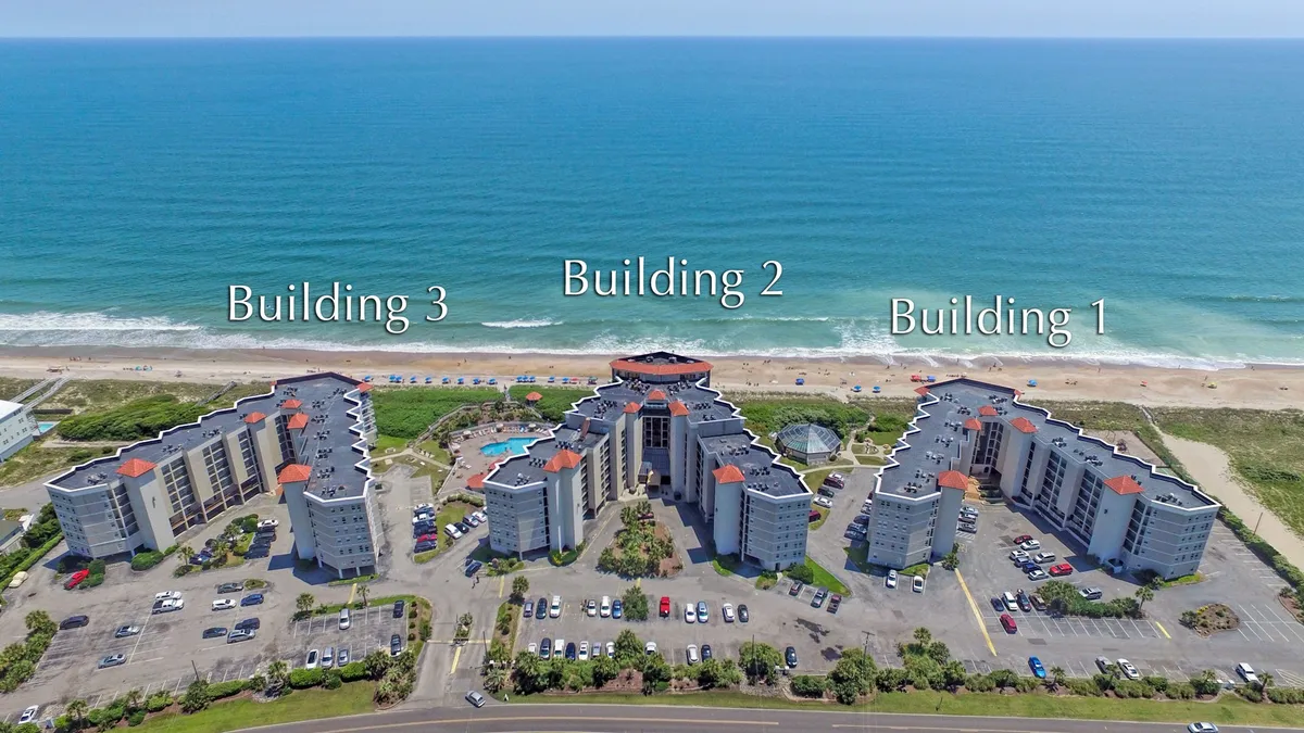 A wide view of the St. Regis resort exterior, featuring three buildings set against the ocean backdrop, a large swimming pool, and the adjacent parking area. The ocean is visible just beyond the property, evoking a sense of direct beach access and resort life.