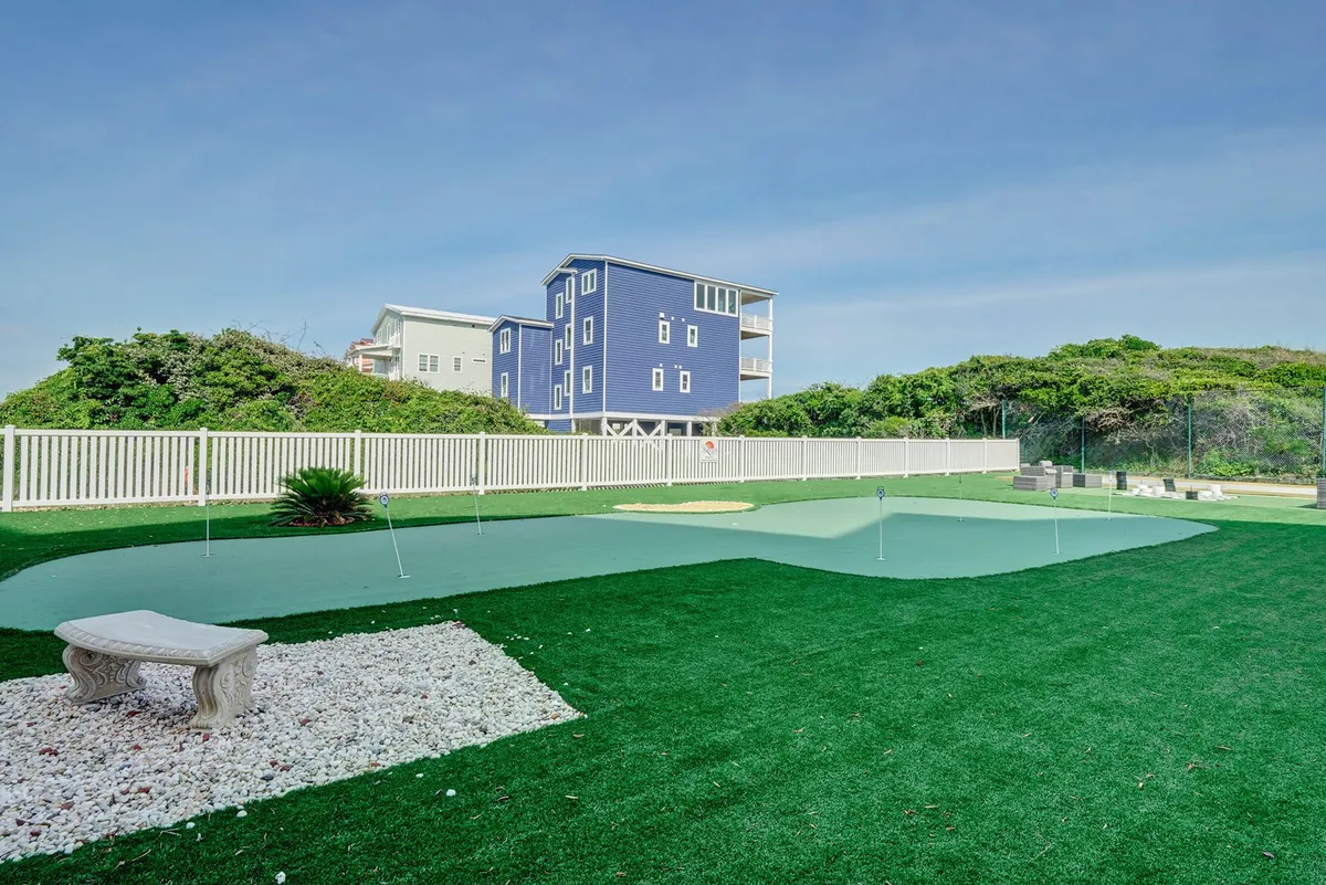 A neatly manicured putting green with lush grass, a white fence, and a blue house in the background. The putting area looks ideal for golf practice or a relaxing game, with space for socializing as well.
