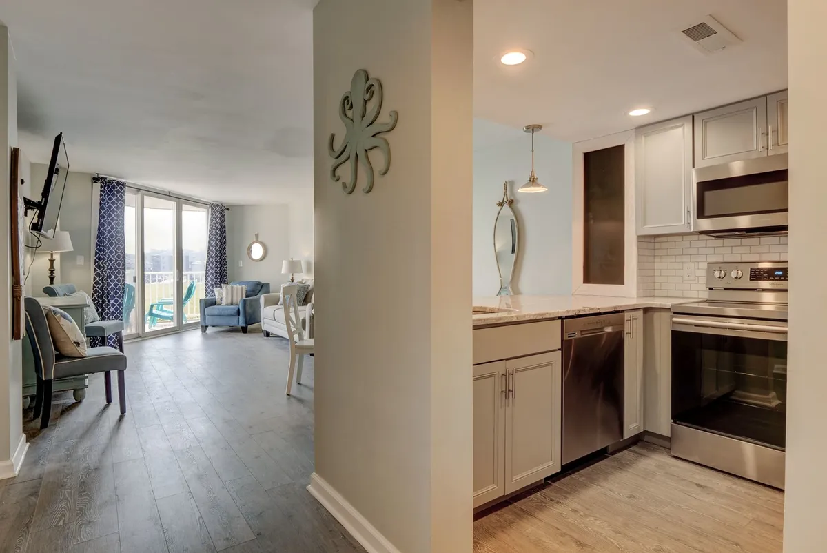 This image highlights a bright, inviting living area with comfortable seating, kitchen appliances in view, whimsical octopus wall decor, and large windows that frame the ocean. The open layout seamlessly connects the living and kitchen spaces, encouraging socializing and relaxation while enjoying the outside scenery.