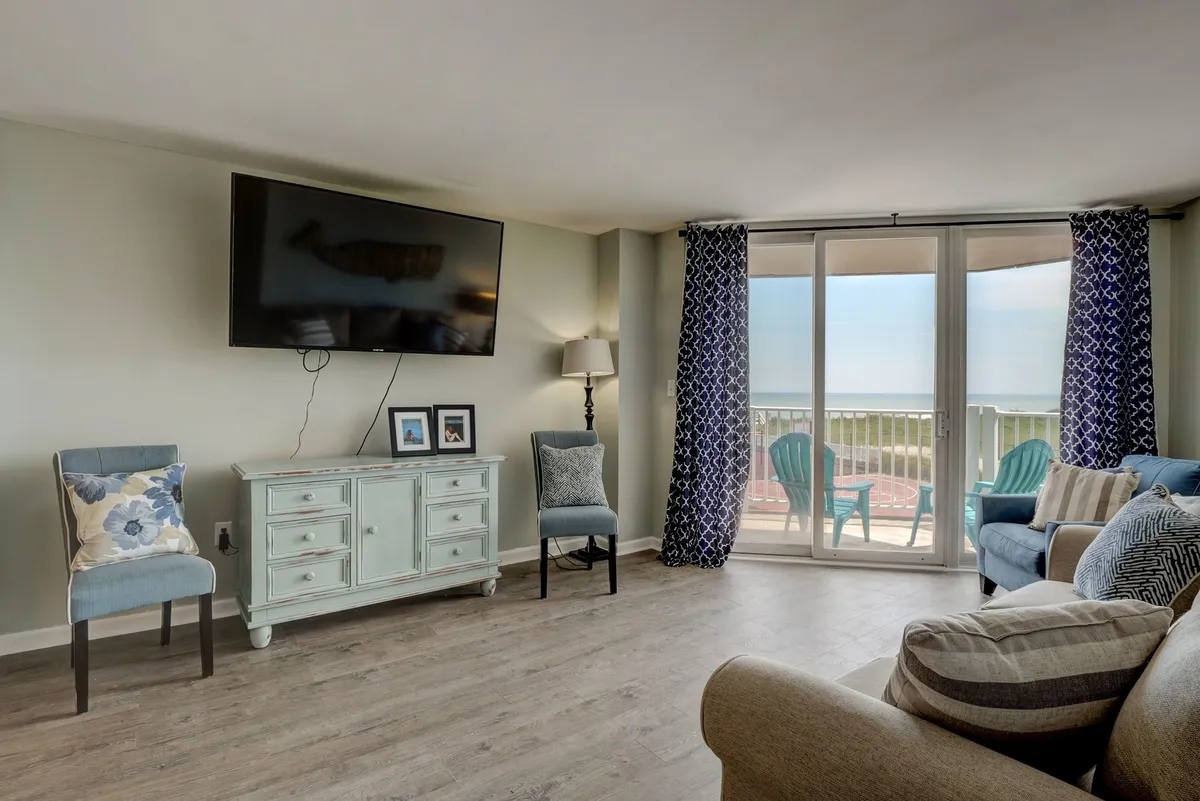 A welcoming living area featuring a comfortable seating arrangement, a light blue sideboard, a TV mounted on the wall, and a large sliding glass door opening to a balcony. The space is bright and relaxed, ideal for family gatherings and easy living.