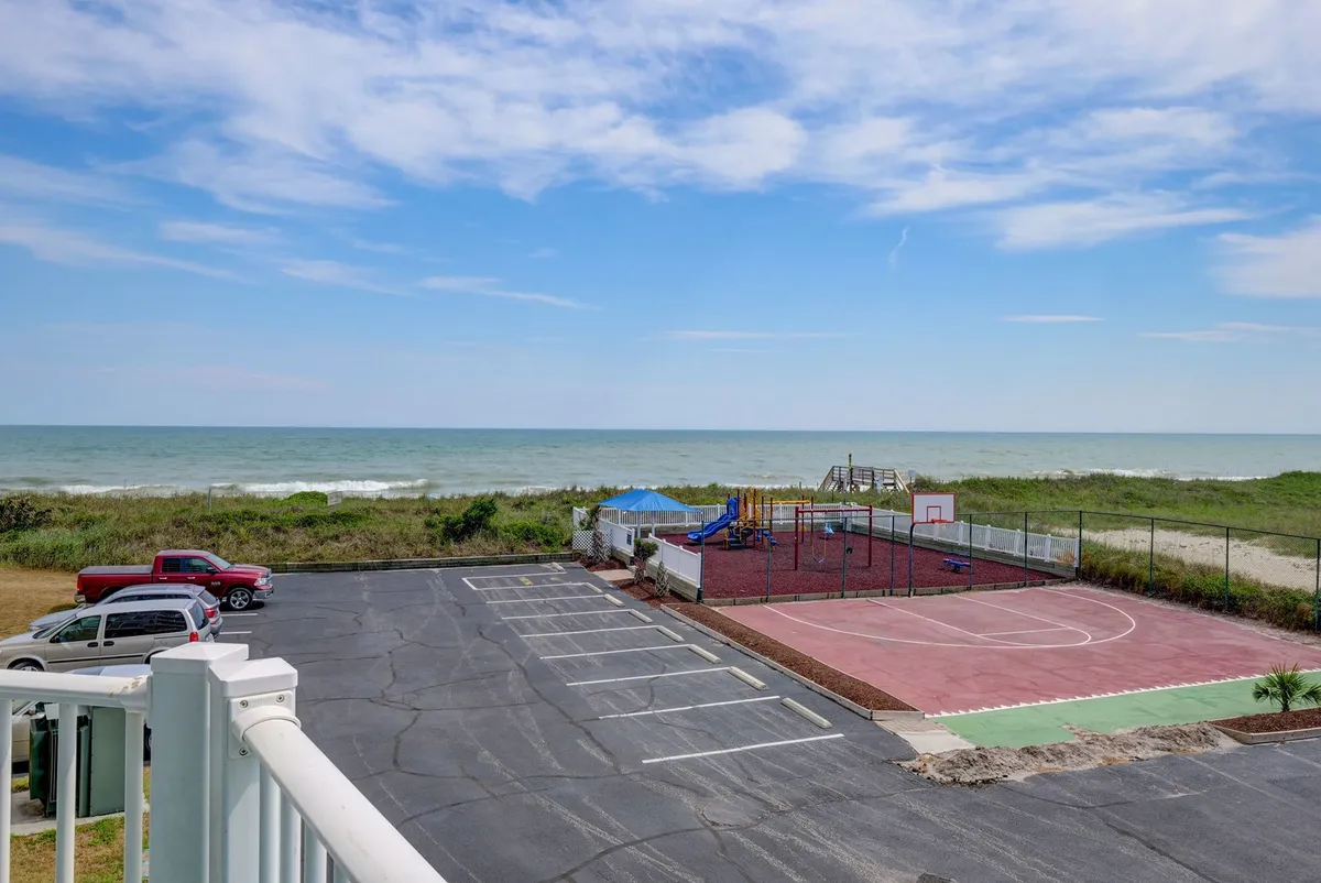 The image showcases the condo's exterior with sweeping ocean views, a vibrant playground area filled with children laughing and playing, and a basketball court where friends and families gather for fun games, all against the backdrop of a stunning sunset. The parking lot is also visible, indicating ample, convenient access for guests.