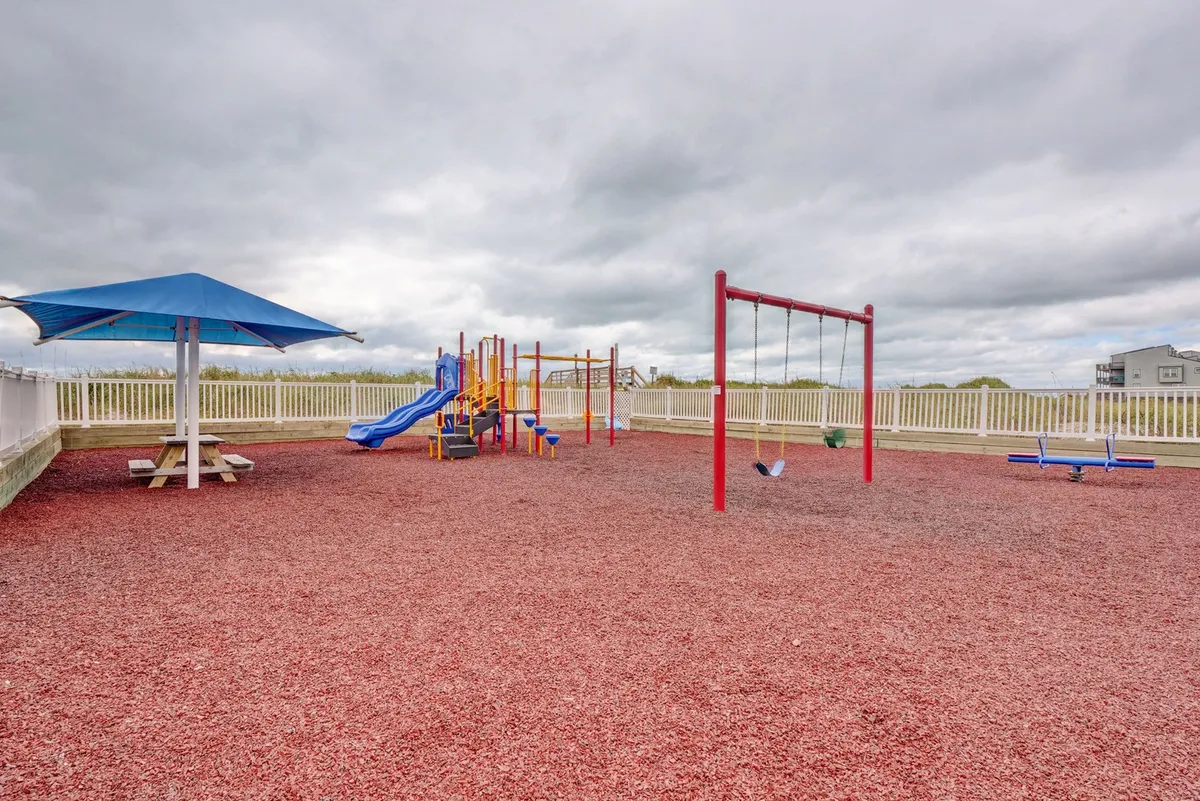 A lively playground with colorful equipment, swings, and a picnic table under a shelter. The setting is designed for children and families to gather and play, with green grass and open space all around.