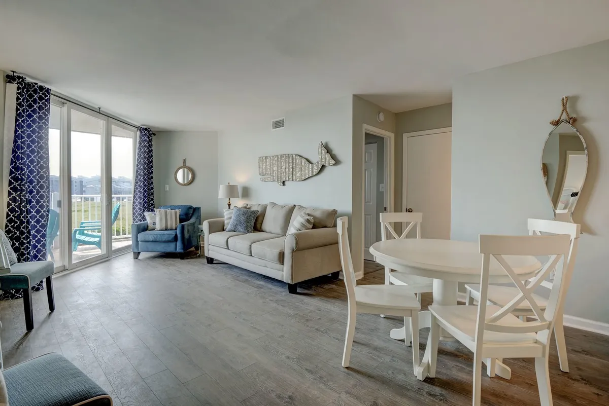A casual, cozy scene shows a sofa, dining table, and wide balcony doors that let in plenty of natural light. Decorative fish wall art adds a coastal touch, and the arrangement suggests a space for relaxed meals, conversations, or game nights with the ocean just beyond.