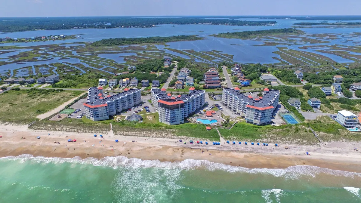 A wide shot of beachfront condos with a large swimming pool in the foreground and the expansive sandy beach and ocean waves just steps away. The atmosphere is relaxed and inviting, showing the seamless blend of condo comfort and outdoor recreation.