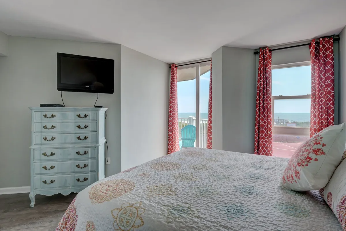 A serene bedroom featuring a bed with a decorative quilt, a blue dresser, a wall-mounted TV, and large windows offering a view. The room is filled with natural light, creating a welcoming and peaceful atmosphere ideal for relaxation and restful sleep.