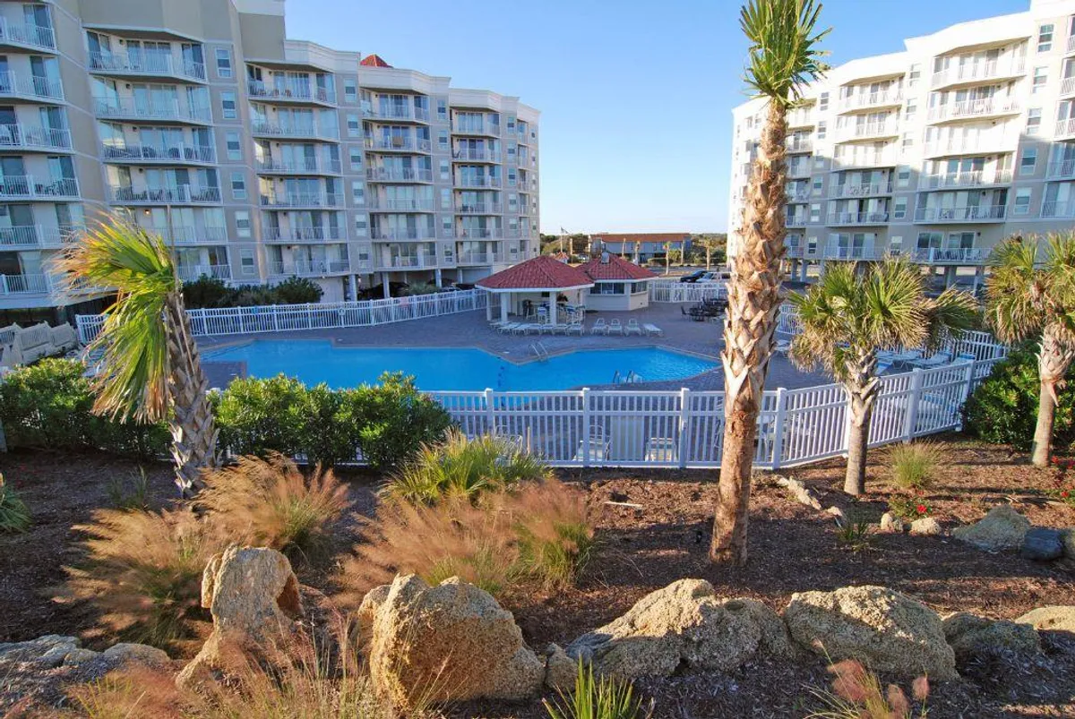 A sparkling swimming pool is surrounded by tall palm trees and several residential buildings. The area looks inviting for both swimming and lounging, with plenty of open sky above, suggesting an outdoor, resort-like atmosphere.