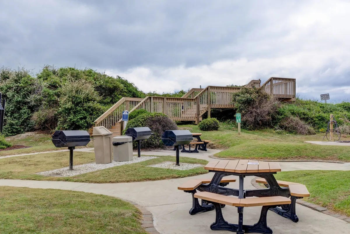 An exterior communal grilling and picnic area with multiple tables, grills, a wooden staircase, and natural surroundings. The setup encourages guests to gather, cook, and eat in a relaxed outdoor setting.